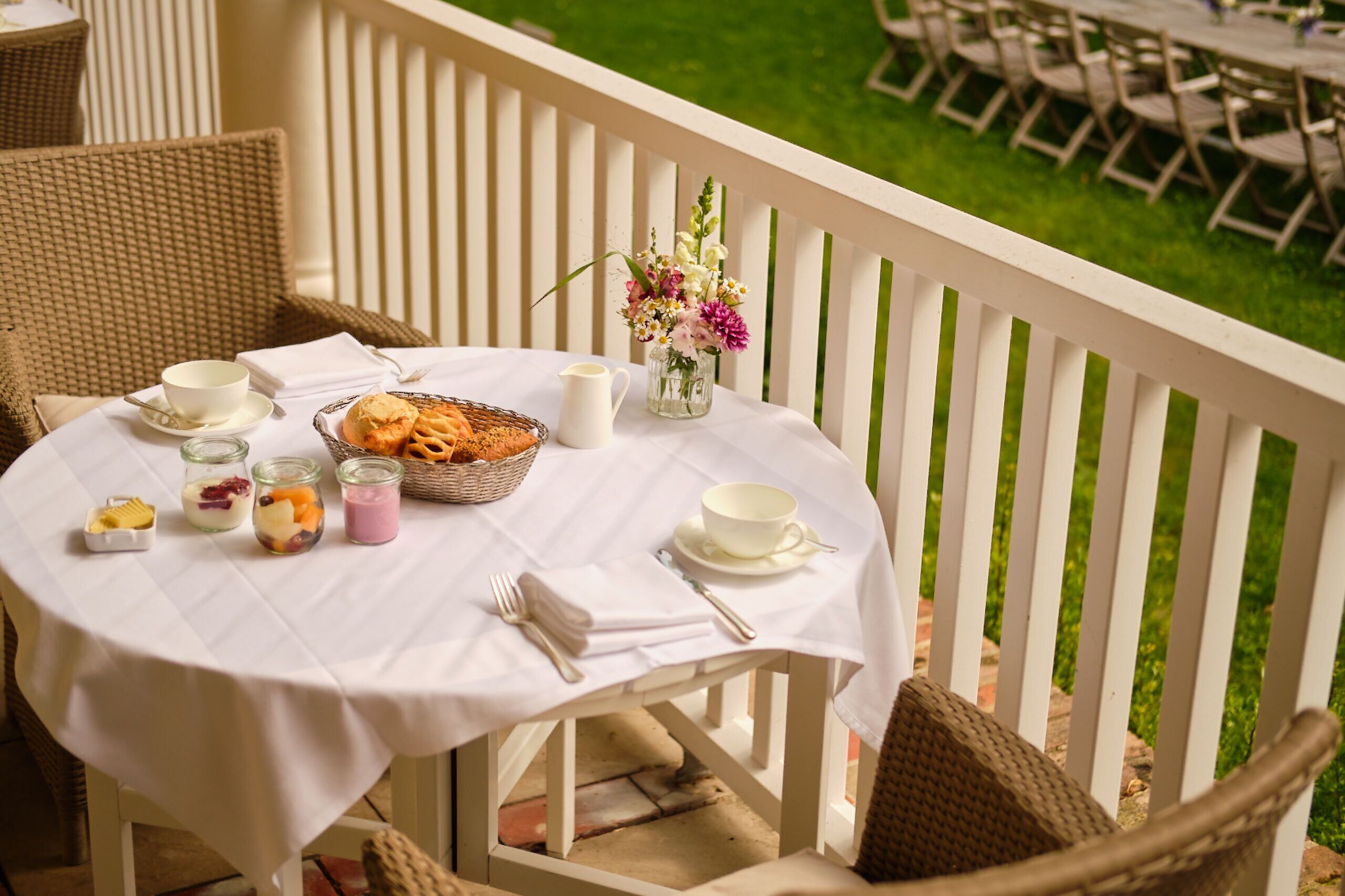 A round table with a white tablecloth, set with a basket of pastries, a pitcher, and a vase of flowers.