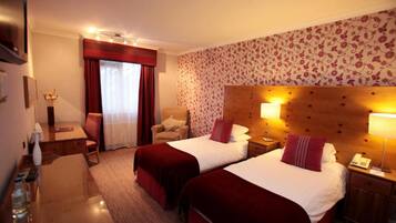 A hotel room with two beds, a desk, a chair, a small table, and a nightstand with a lamp.
