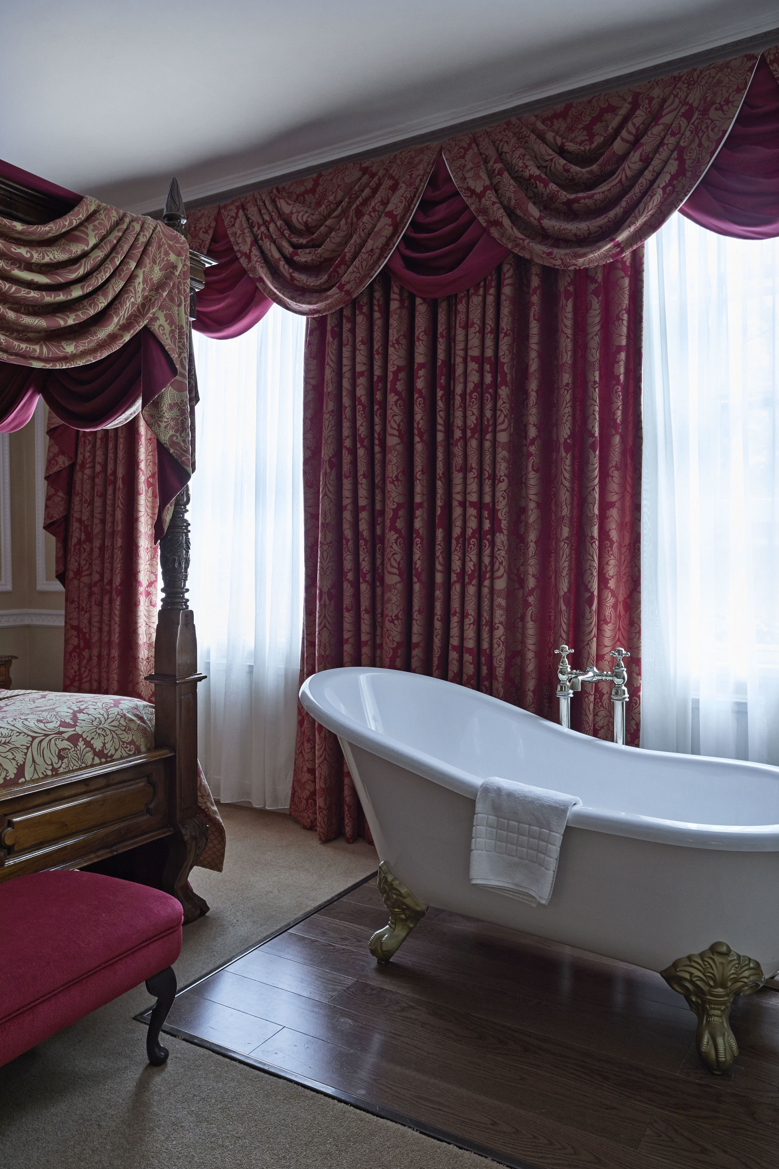 A bathroom with a freestanding bathtub, a wooden bench, and a large window with red curtains.