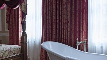 A bathroom with a freestanding bathtub, a wooden bench, and a large window with red curtains.