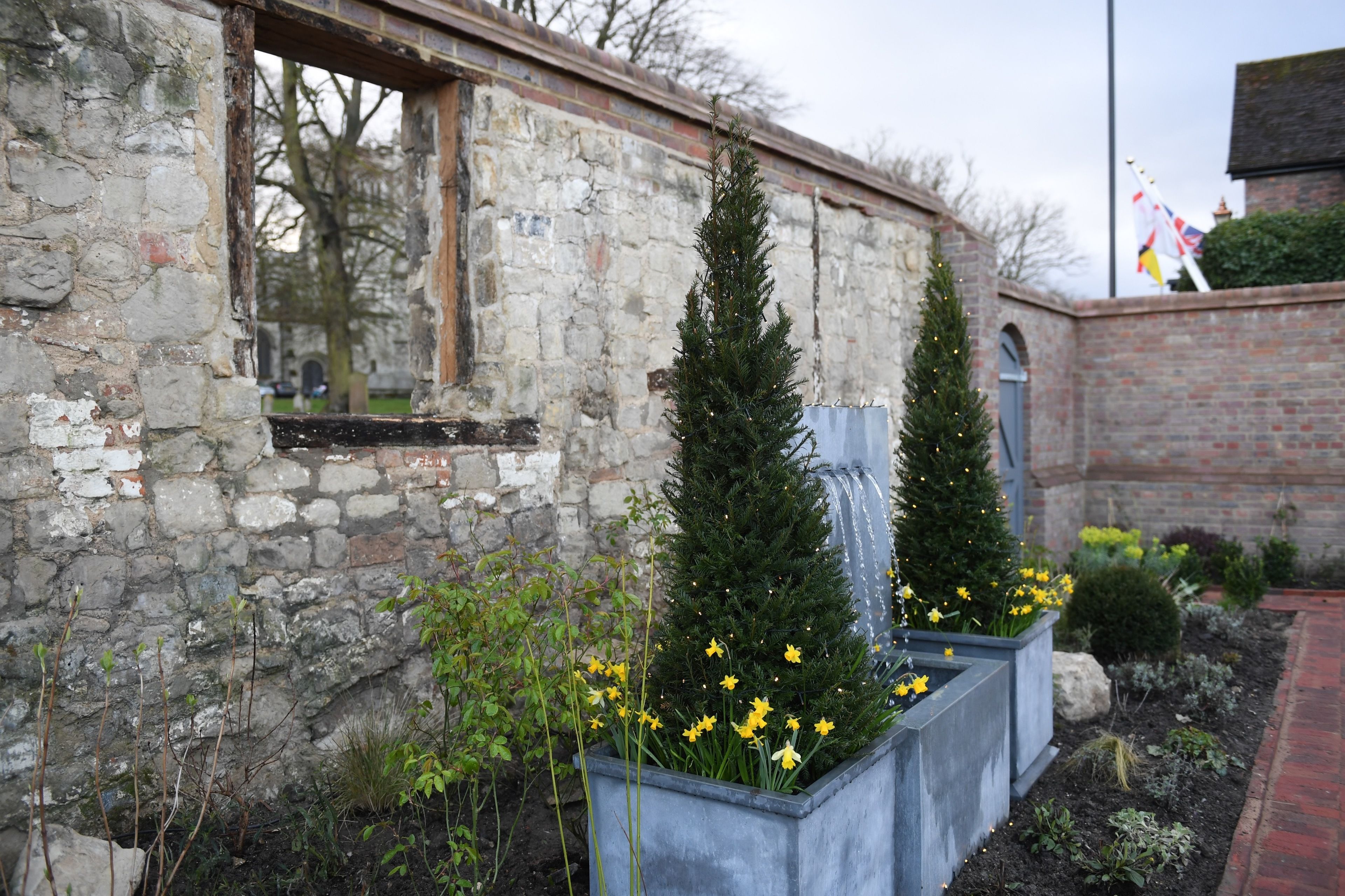 Un edificio de piedra con una ventana y un muro de ladrillo. Se ven dos árboles de hoja perenne y macizos de flores amarillas.