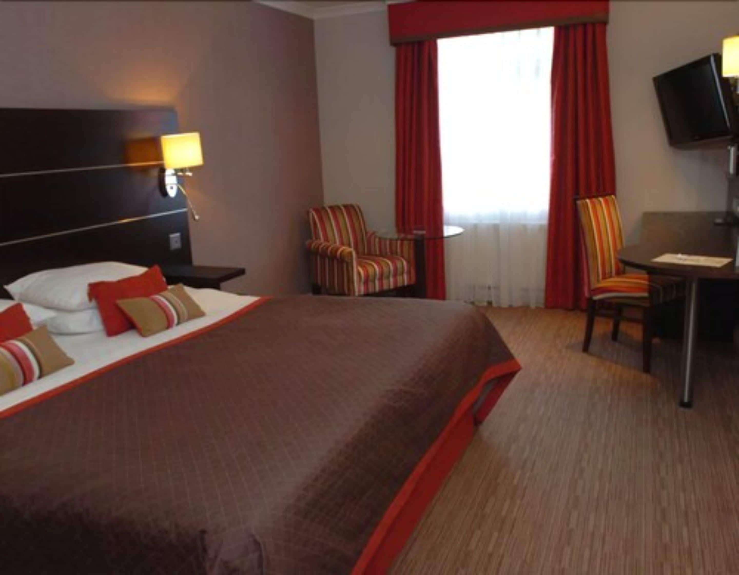 Een hotelkamer met een groot bed, een bureau, twee stoelen, een televisie en een raam met gordijnen.