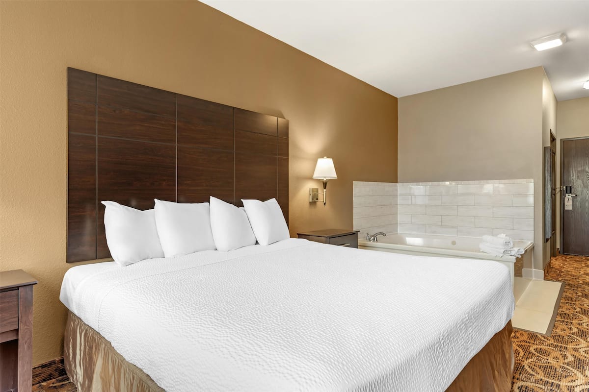 Ein Hotelzimmer mit einem großen Bett, einem Nachttisch, einer Wandleuchte, einem Badezimmer mit einer Badewanne und einer Tür.