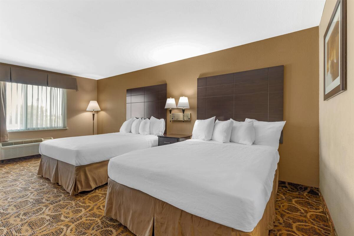 Ein Hotelzimmer mit zwei Betten, einem großen Fenster mit Vorhängen, einer Stehlampe und einem gerahmten Bild an der Wand.