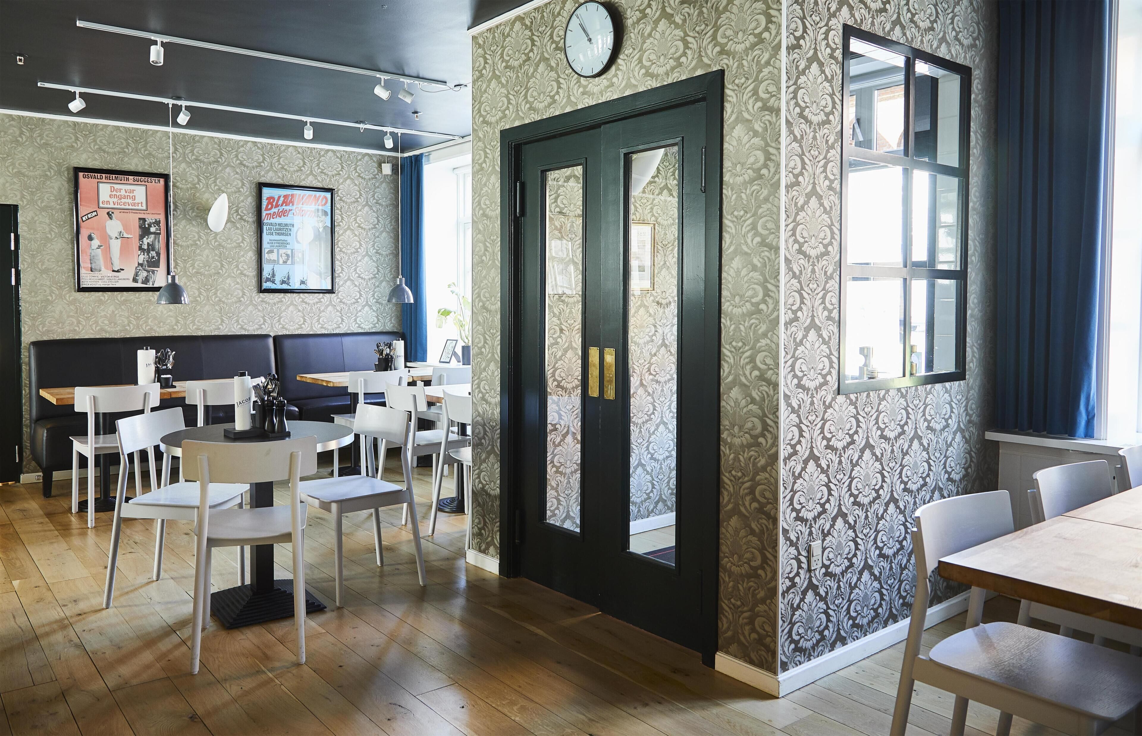 A restaurant with a modern interior, featuring a wooden floor, white chairs, a black door, and patterned wallpaper.