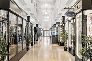 Un pasillo con tiendas de escaparate de vidrio, incluyendo una joyería y una tienda de hotel, con plantas en macetas y suelo de baldosas.