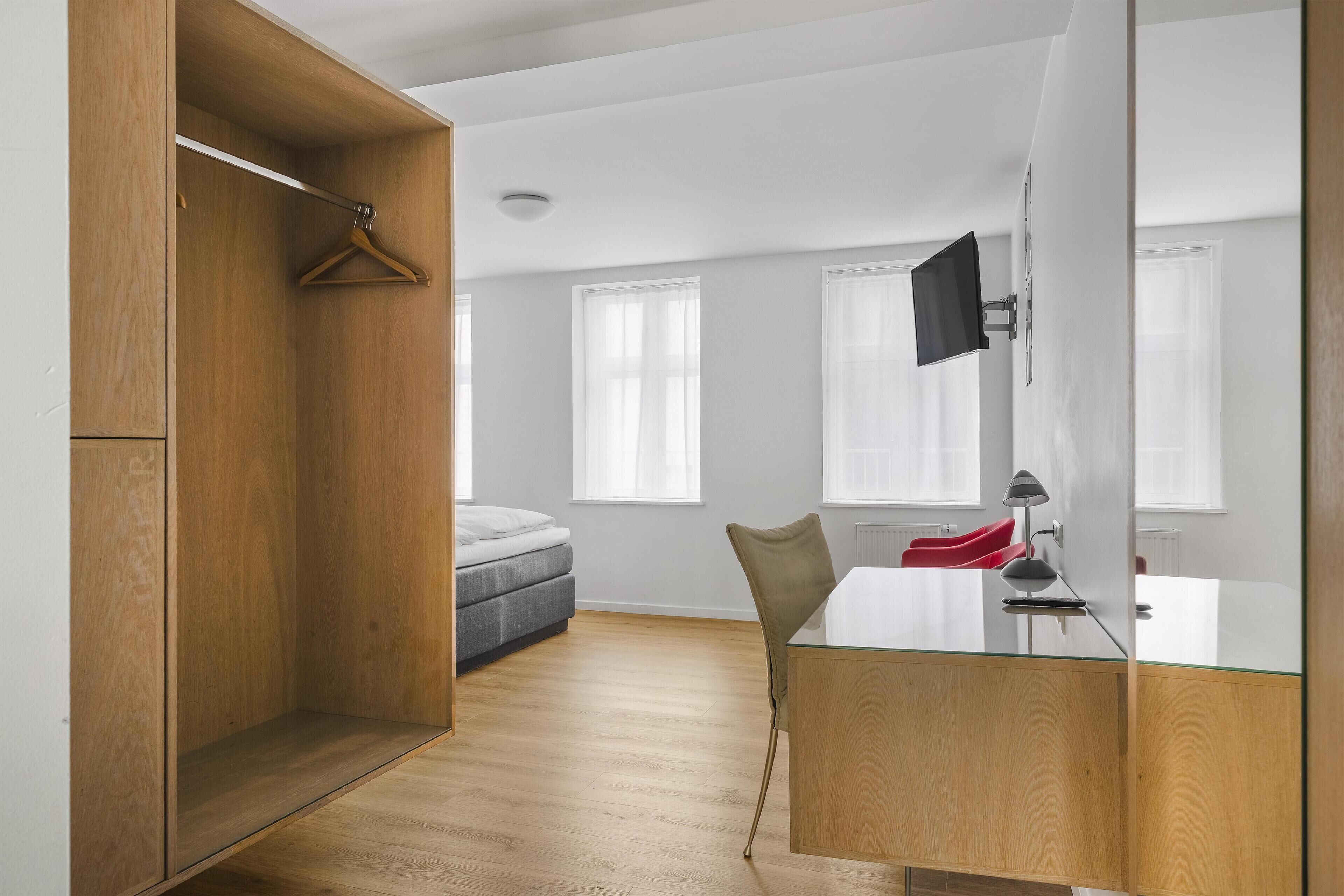 A room with a bed, a desk with a chair, a wooden wardrobe, and a TV mounted on the wall.
