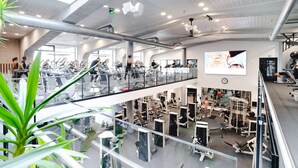 Un gimnasio de varios niveles con diversos equipos de ejercicio, una pantalla grande que muestra información de entrenamiento y una barandilla de vidrio con vistas al nivel inferior.