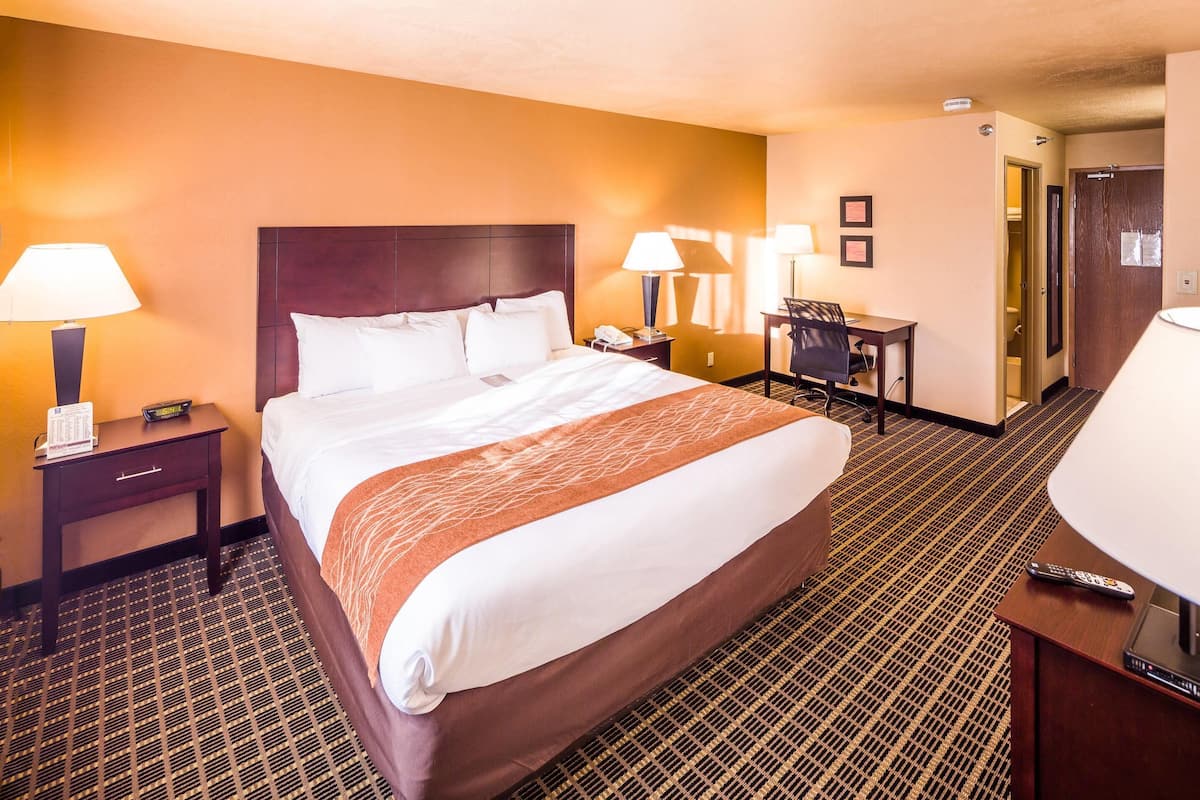 Habitación de hotel con una cama grande, dos mesitas de noche con lámparas, un escritorio con silla y una puerta que da a otra habitación.