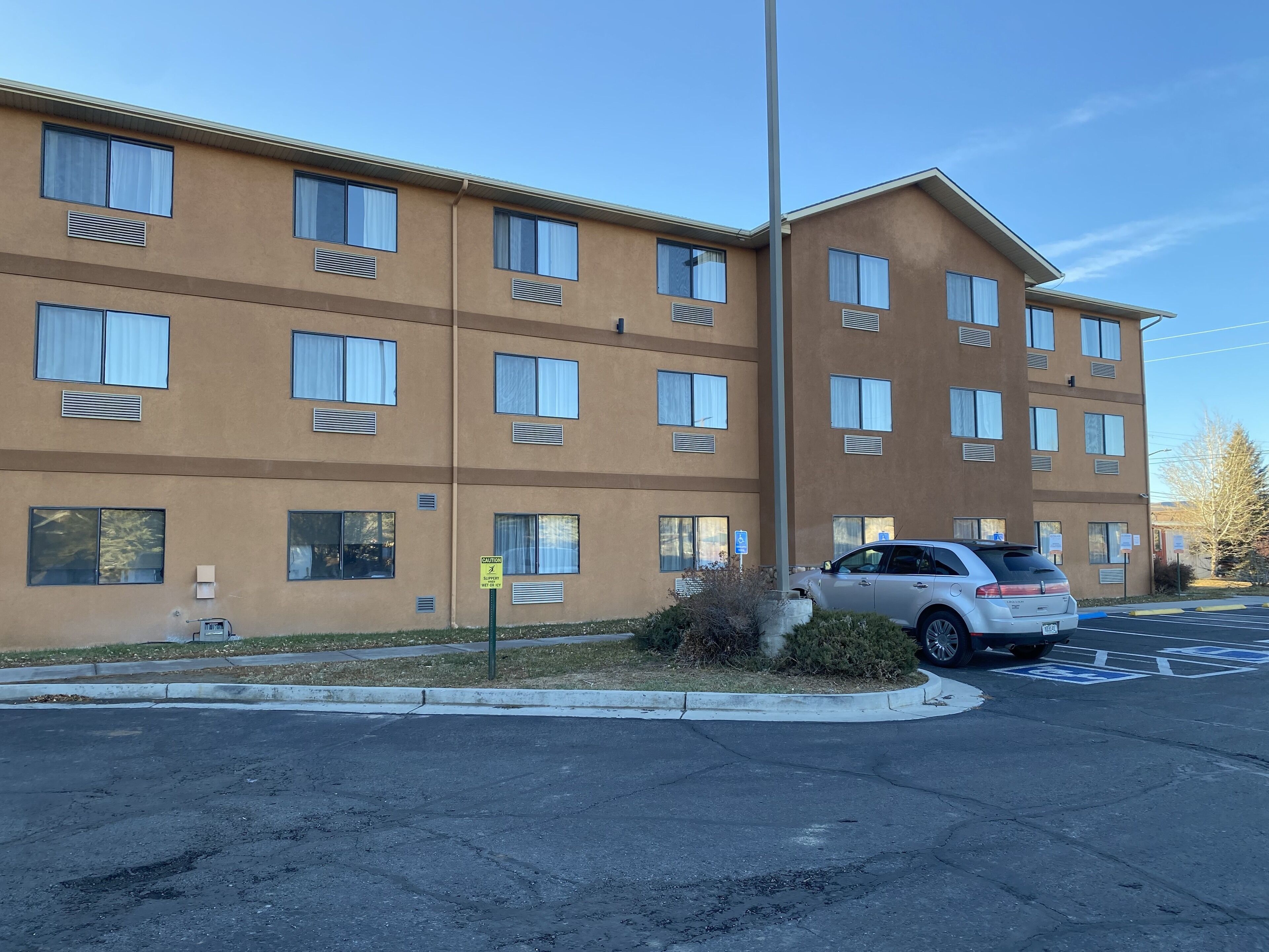 Un hotel de varios pisos con fachada color crema, grandes ventanales y un estacionamiento con un coche blanco.