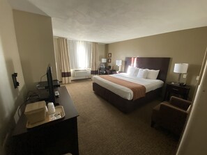 A hotel room with a large bed, a desk with a computer, a chair, and a bedside table with a lamp.