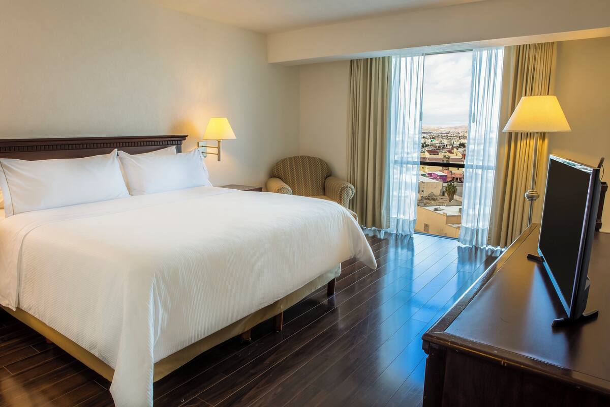 A hotel room with a large bed, two bedside lamps, a chair, a desk with a TV, and a balcony with a view of the city.