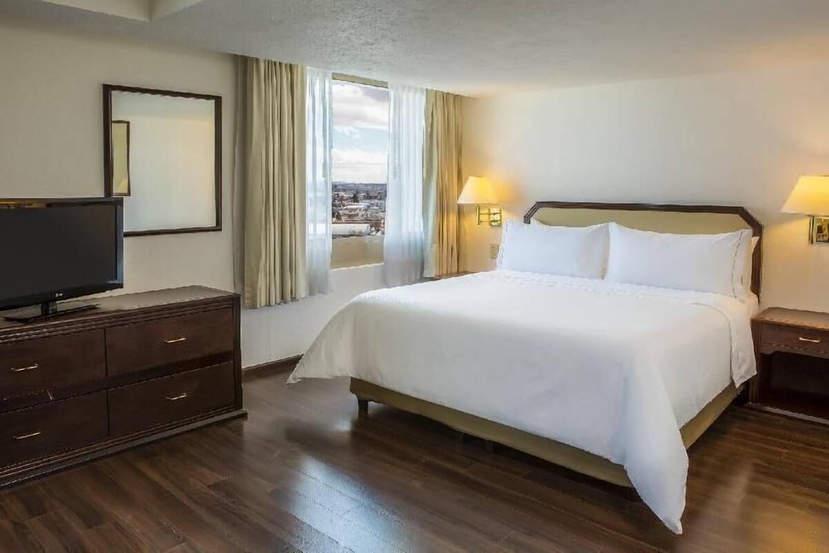 A hotel room with a large bed, a television on a dresser, and a view of the city through the window.