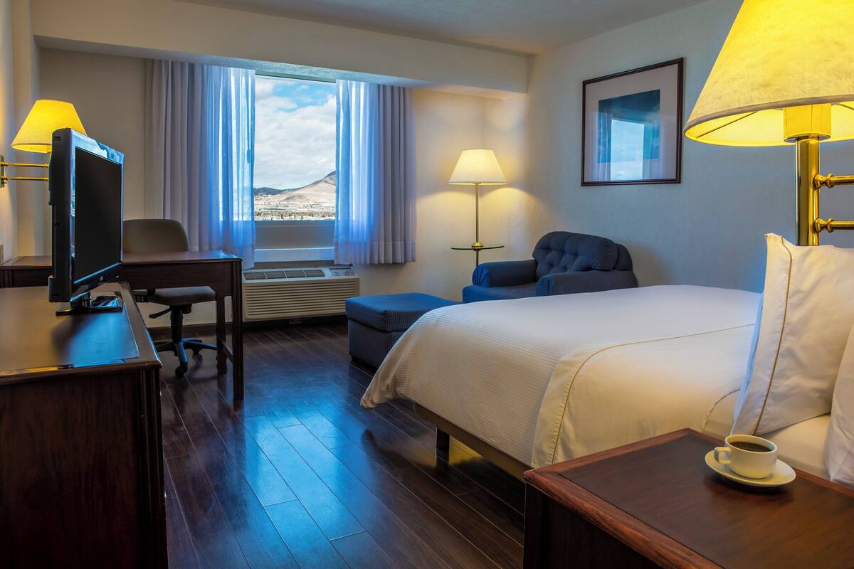 A hotel room with a bed, a desk with a computer, a chair, a sofa, a lamp, and a window with curtains.