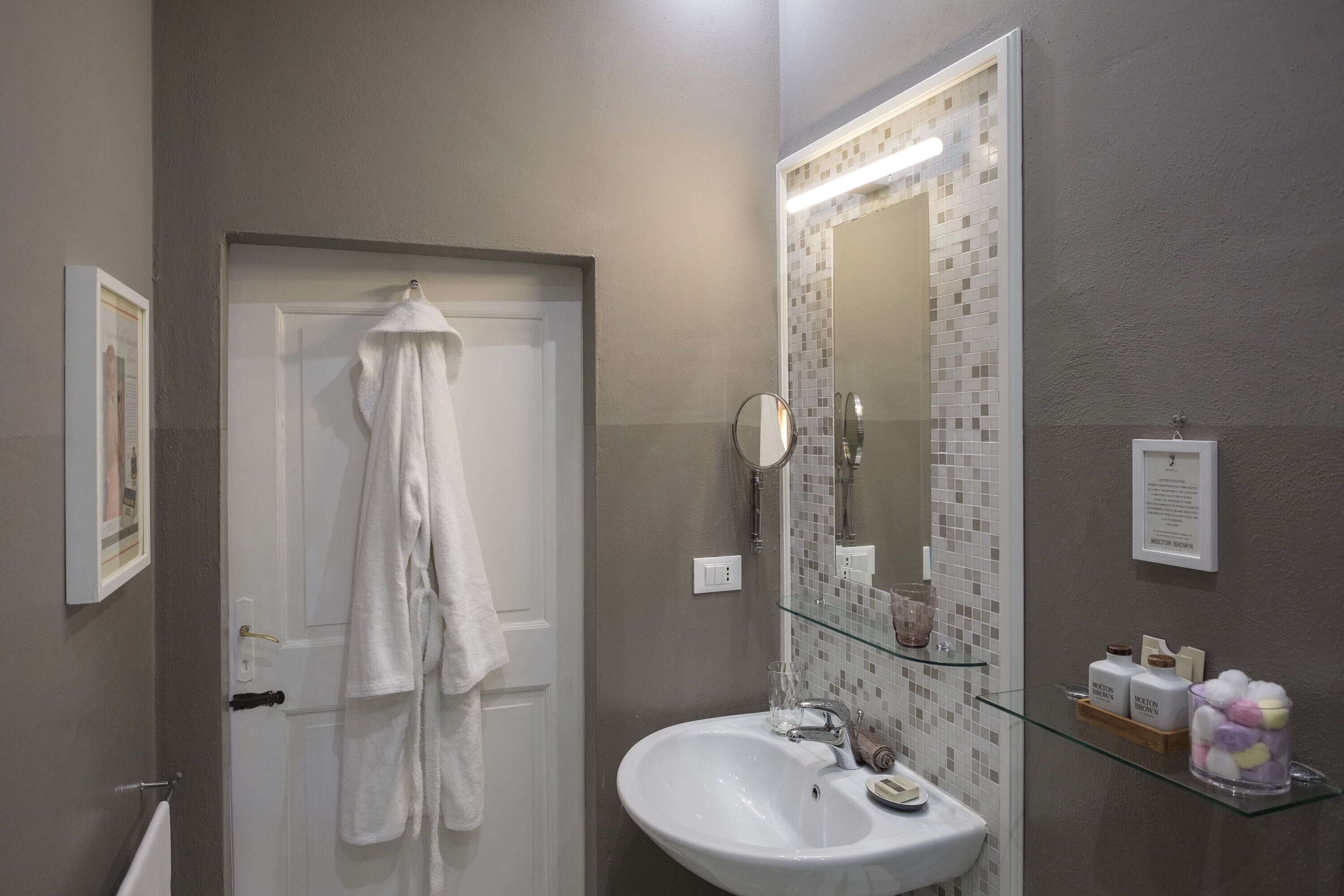 Un bagno con un lavandino bianco, uno scaffale in vetro con articoli da toilette, uno specchio e un asciugamano appeso alla porta.