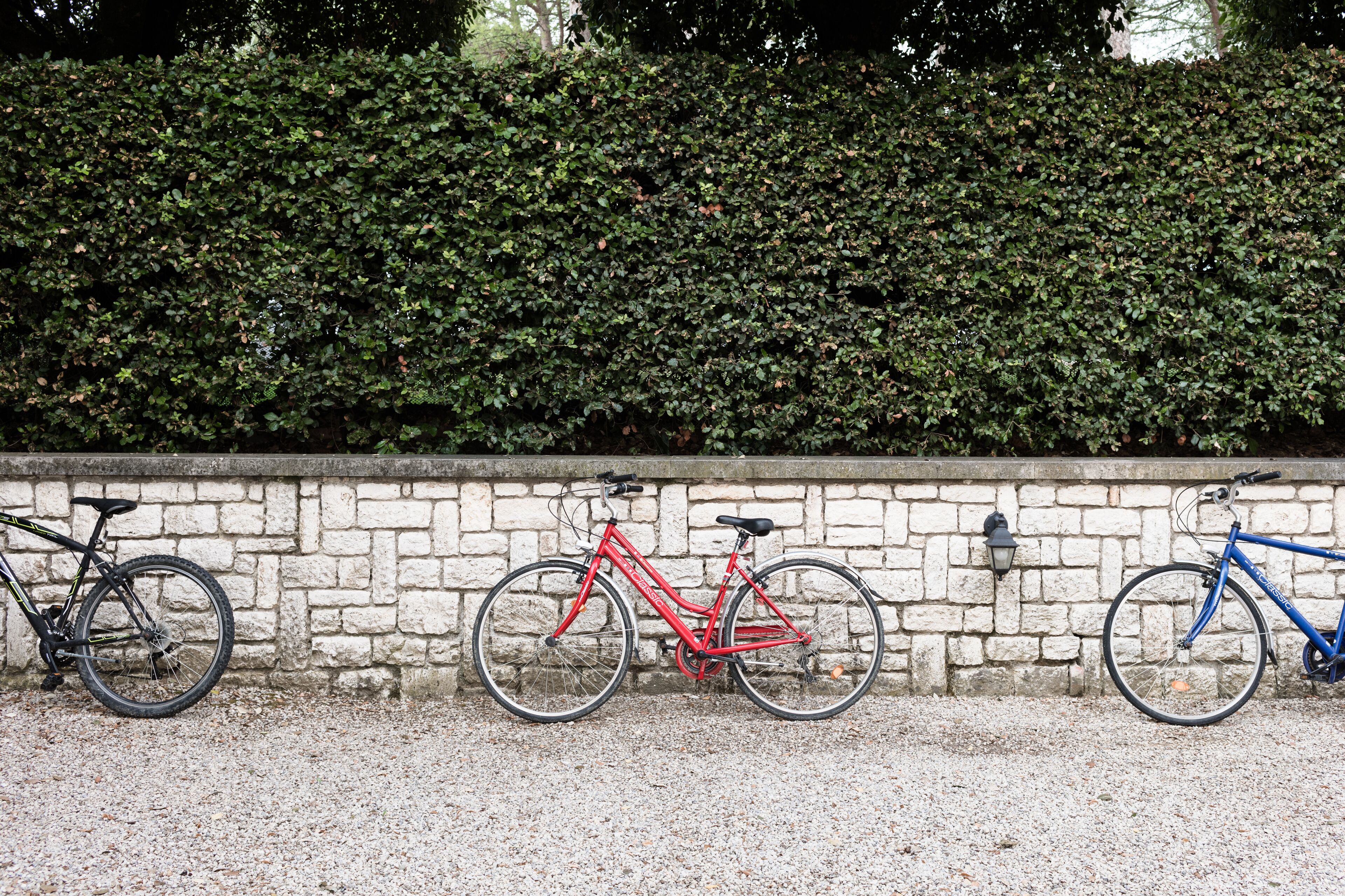 Three bicycles parked against a stone wall with a hedge in the background.