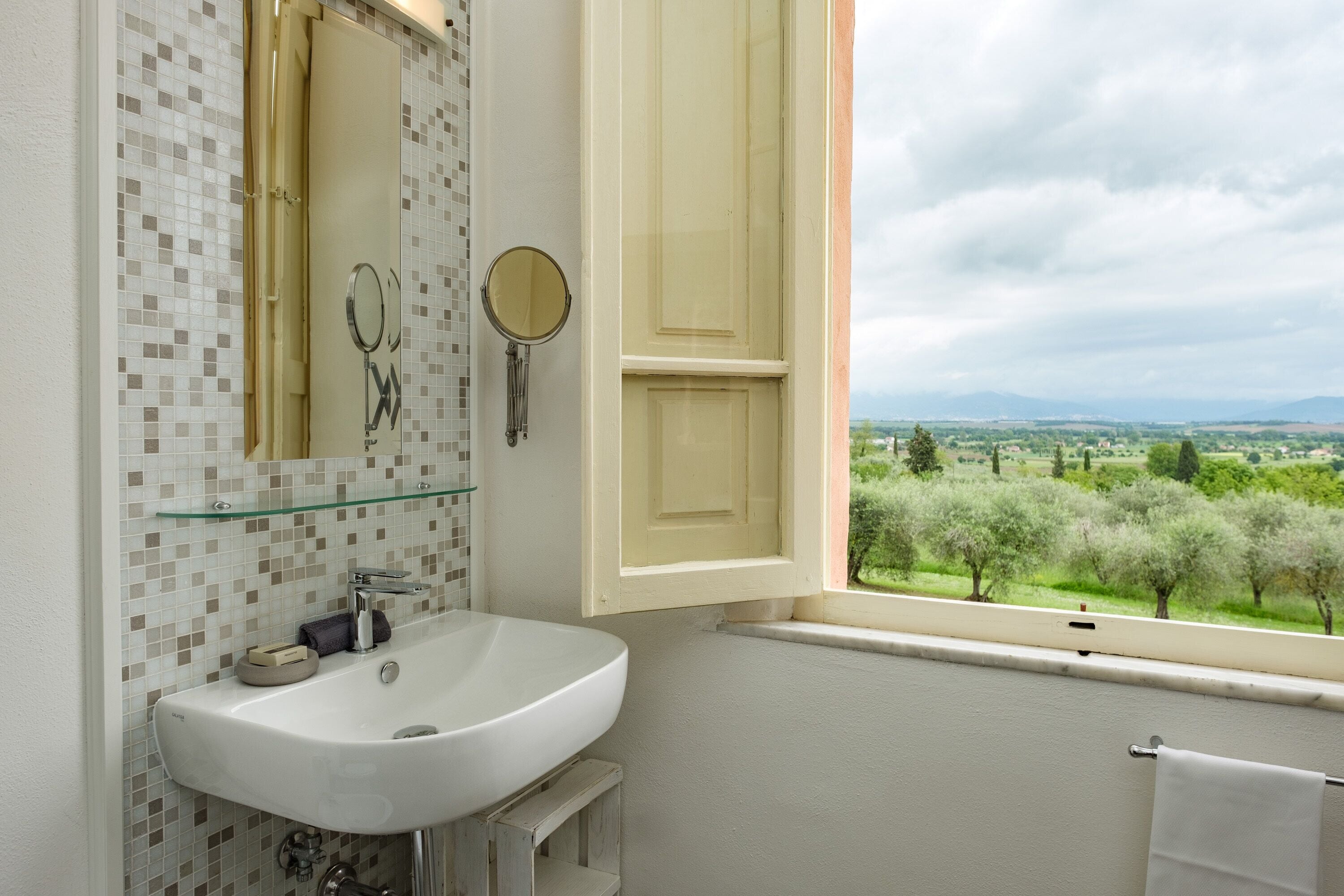 A bathroom with a white sink, two mirrors, and a window with a view of green trees and hills.