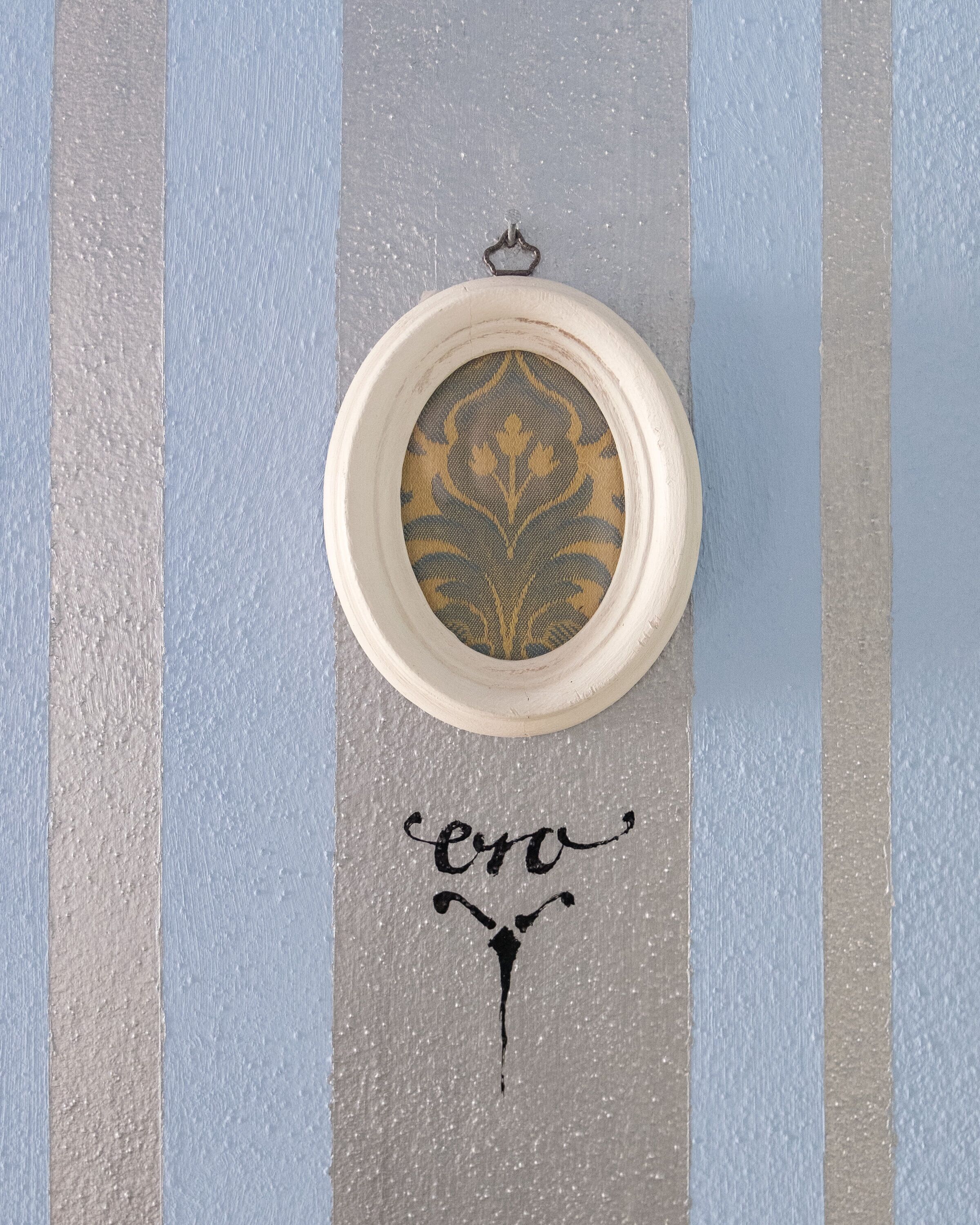 Un objet circulaire à la surface texturée, muni d’une chaîne pour le suspendre, placé sur une surface à rayures ornée d’un motif décoratif, avec le mot « ero » écrit en dessous.