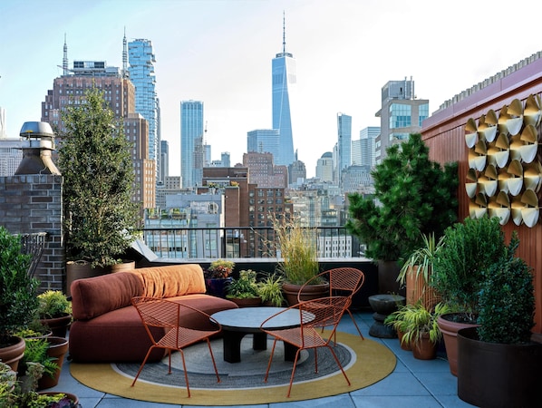 A rooftop terrace with a sofa, chairs, and potted plants, overlooking a city skyline.