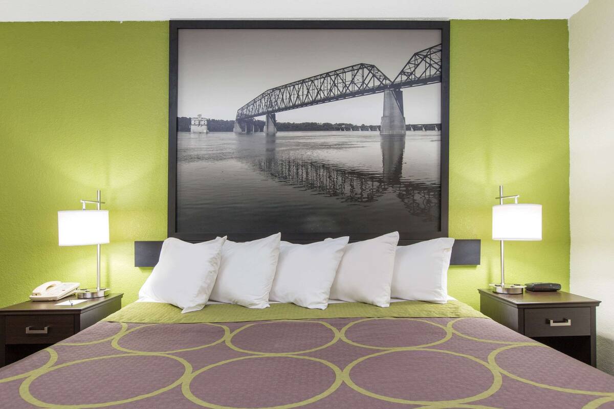 Une chambre d’hôtel avec un lit, deux tables de chevet, une petite table, une lampe et un grand tableau encadré représentant un pont, accroché au mur.