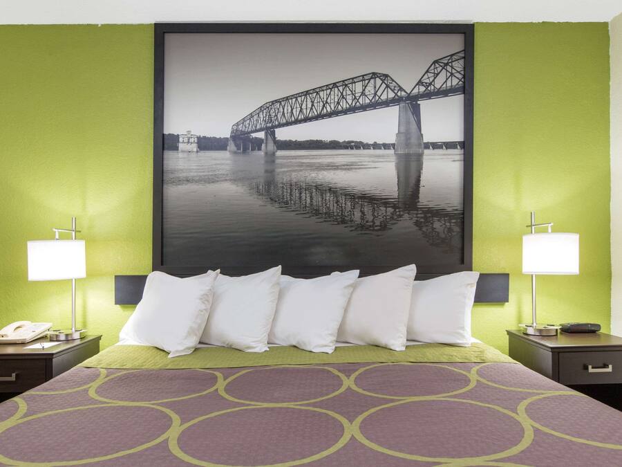 Una habitación de hotel con cama, dos mesitas de noche, una lámpara y un cuadro grande con un puente en la pared.