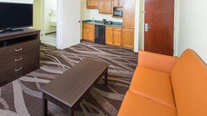 A compact hotel room with a sofa, a TV on a wooden cabinet, a kitchenette with wooden cabinets, a microwave, and a refrigerator.