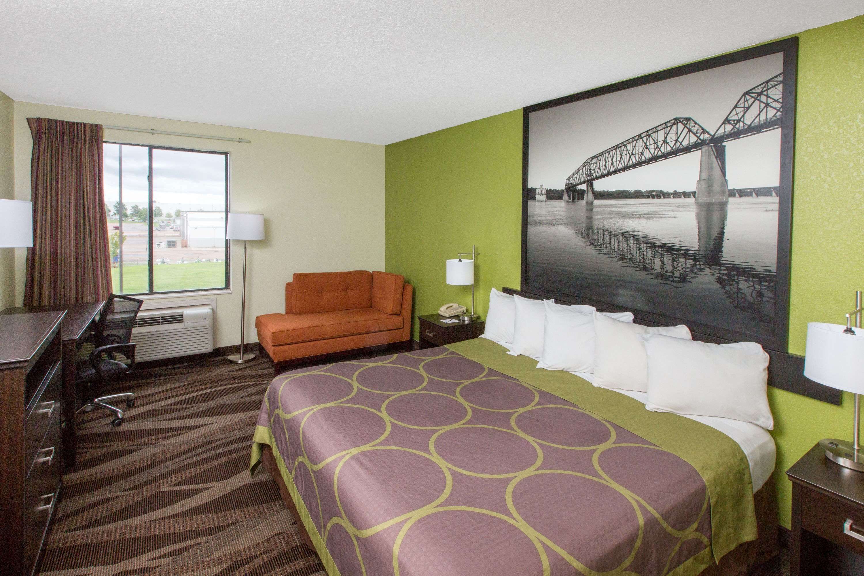 Una habitación de hotel con una cama grande, un escritorio con silla, un sofá pequeño naranja, una ventana con cortinas y un cuadro de un puente.