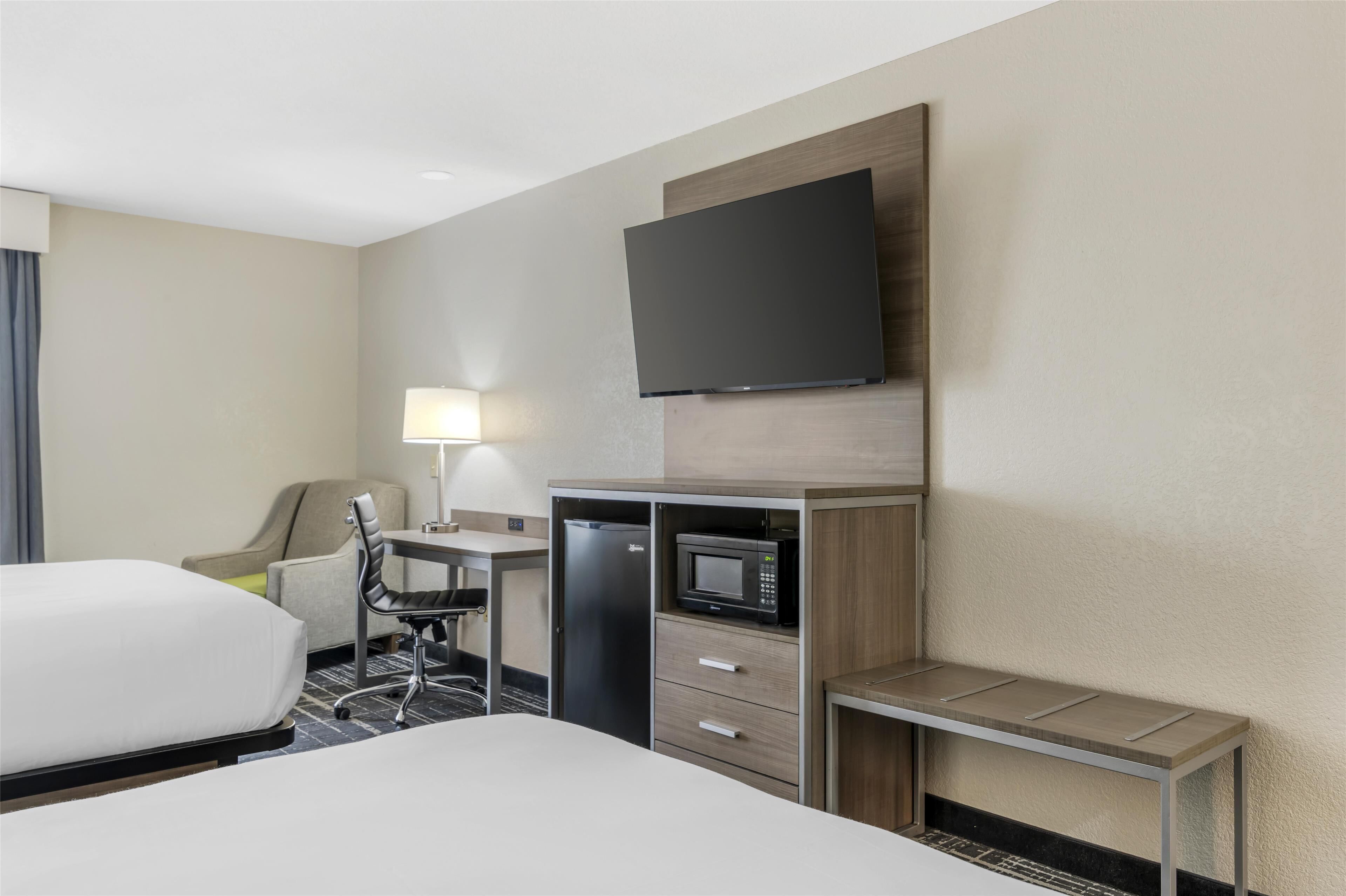 A hotel room with a bed, a desk with a chair, a microwave, a refrigerator, and a flat-screen TV mounted on the wall.