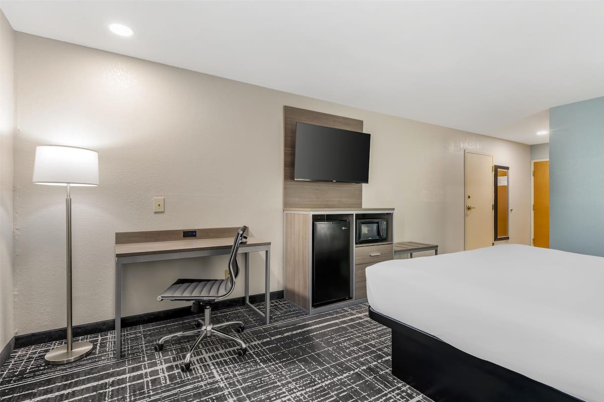 A hotel room with a bed, desk, chair, flat-screen TV, refrigerator, microwave, and a lamp.