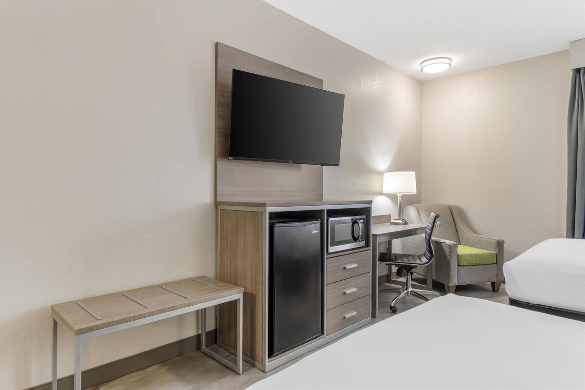 A hotel room with a bed, a desk with a chair, a television mounted on the wall, a refrigerator, and a bench.