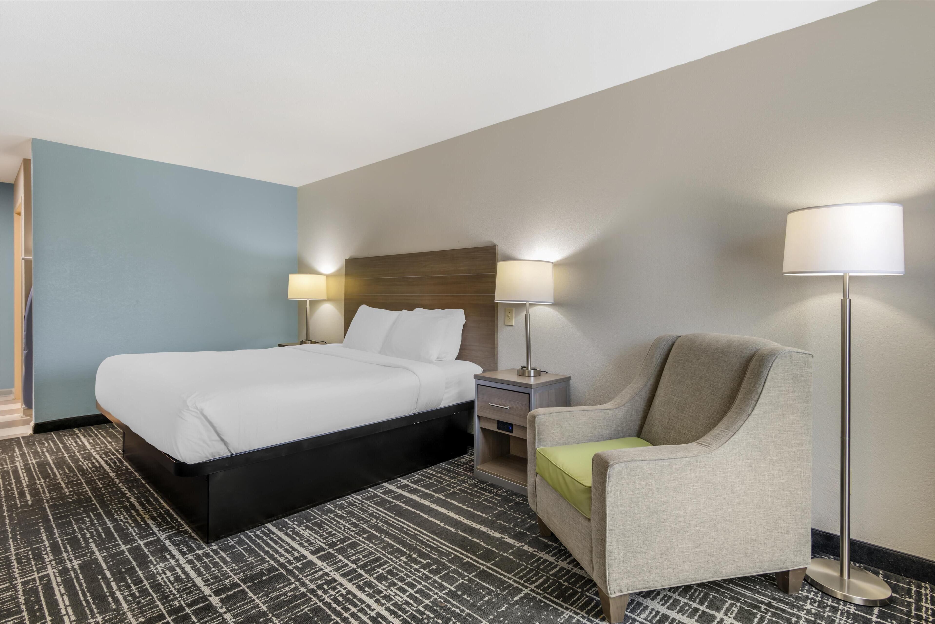 A hotel room with a bed, a chair, a nightstand, and two lamps.