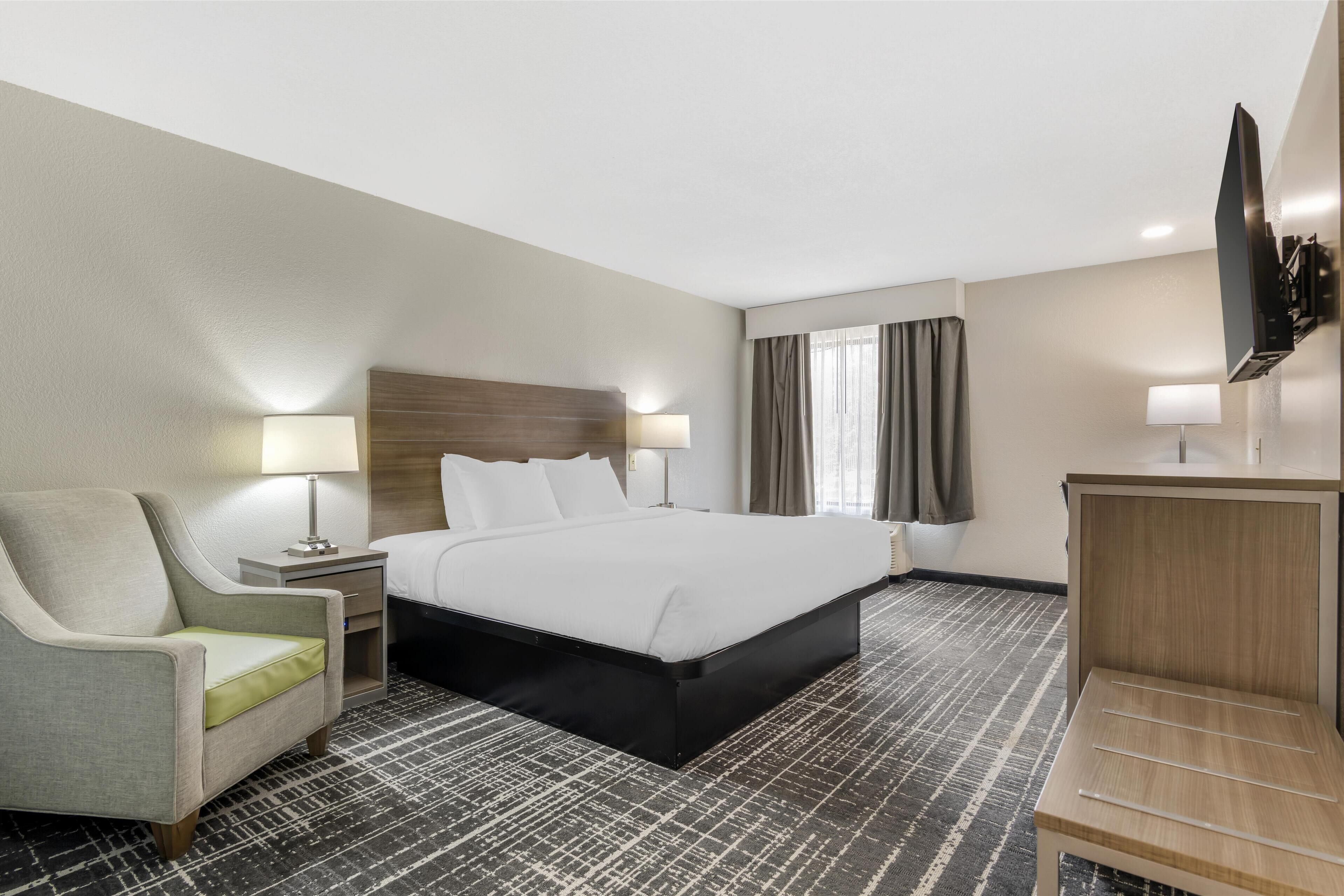 Habitación de hotel con cama, una silla, un escritorio, un televisor y ventana con cortinas.