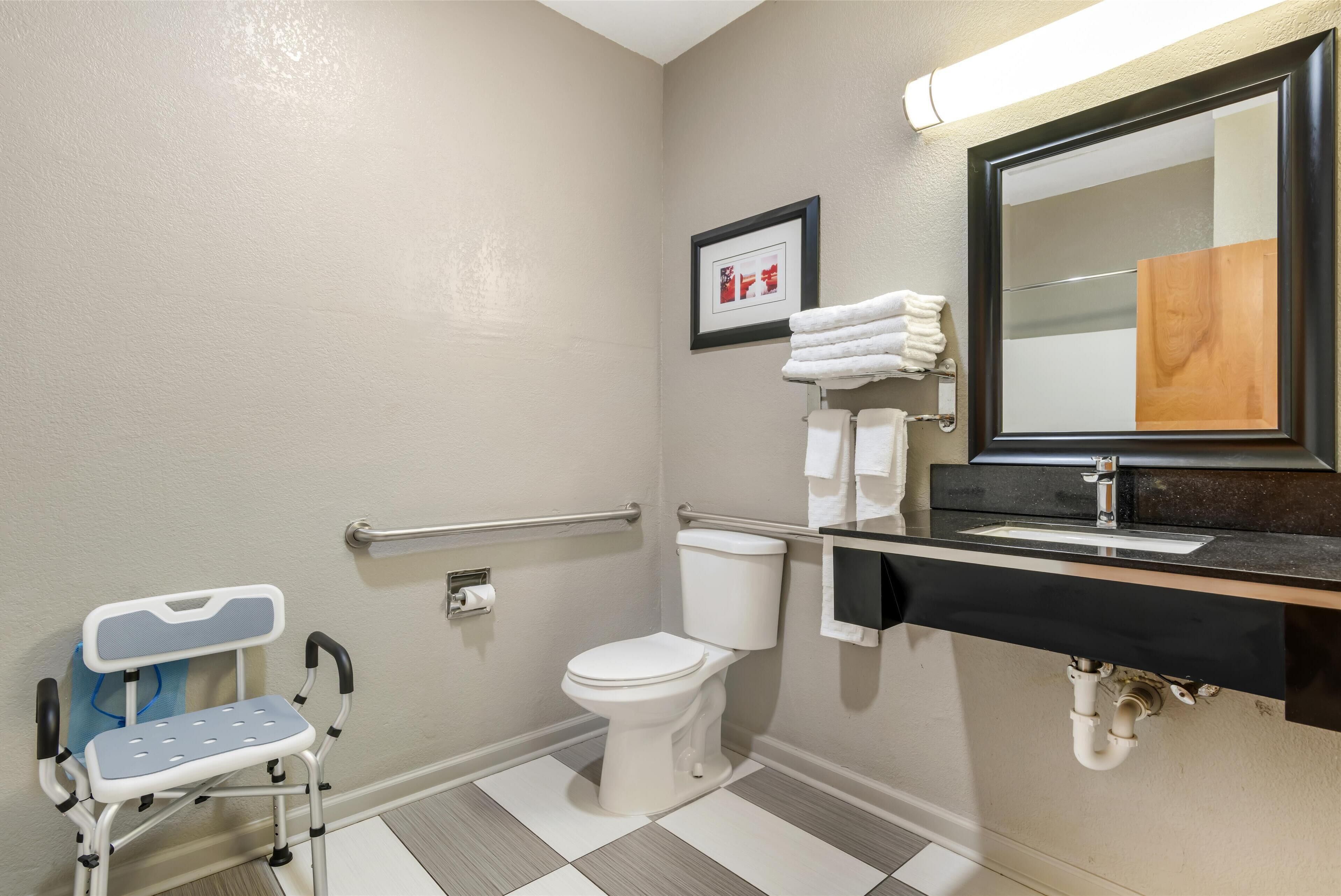 A bathroom with a white toilet, a sink with a black countertop, a framed picture on the wall, and a wheelchair.