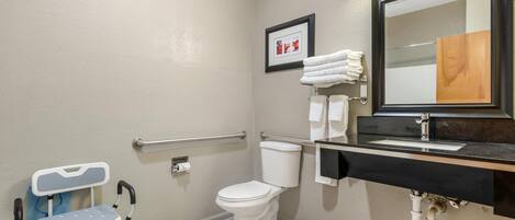 A bathroom with a white toilet, a sink with a black countertop, a framed picture on the wall, and a wheelchair.