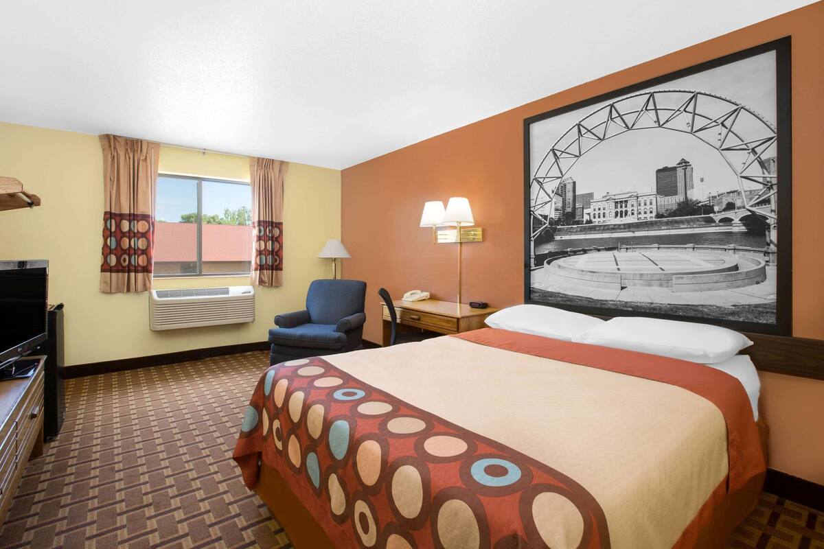 A hotel room with a bed, a desk, a chair, an air conditioning unit, a television, and a large framed picture on the wall.