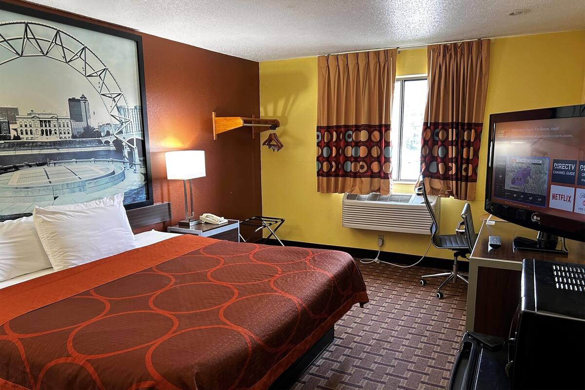 A hotel room with a bed, a desk with a computer, an air conditioning unit, and a large window with curtains.