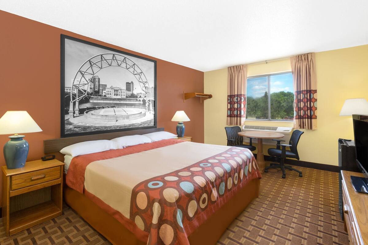 A hotel room with a bed, two bedside tables with lamps, a desk with a chair, a television, and a large window with curtains.