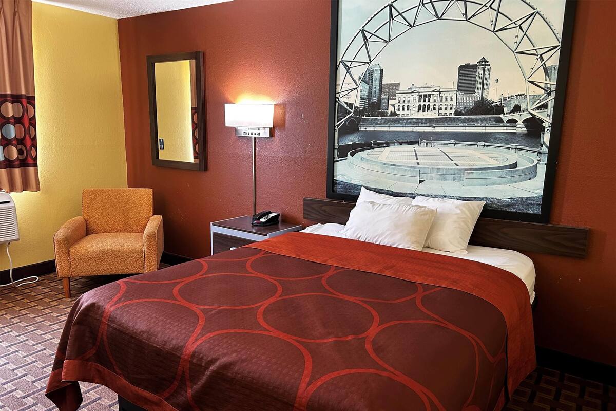 A hotel room with a bed, a chair, a nightstand with a phone, a wall-mounted lamp, and a large cityscape painting.