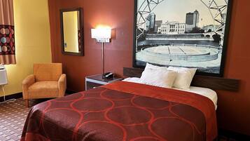 A hotel room with a bed, a chair, a nightstand with a phone, a wall-mounted lamp, and a large cityscape painting.