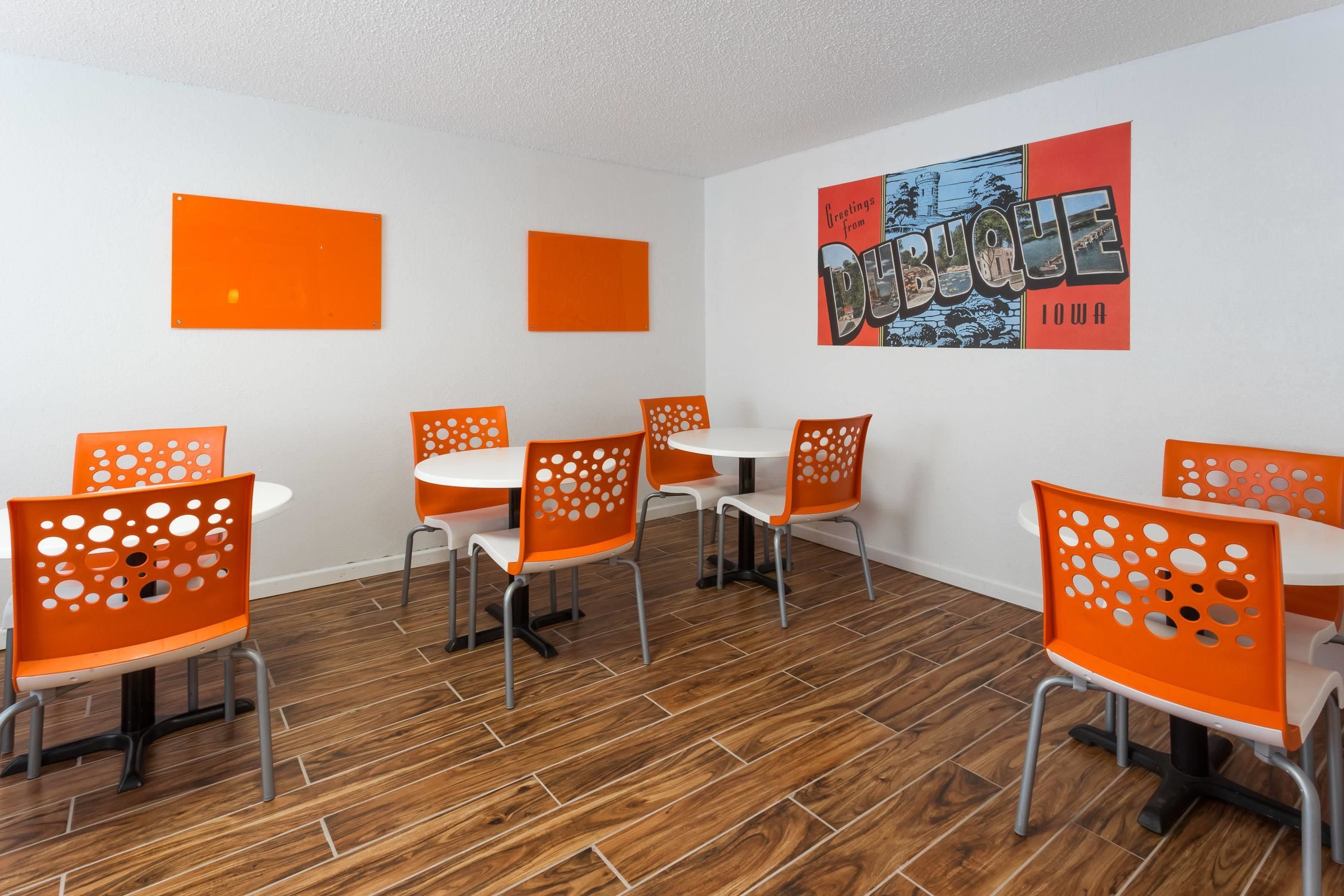 A dining area with orange chairs and white tables, a wooden floor, and a 'Greetings from Des Moines Iowa' poster on the wall.