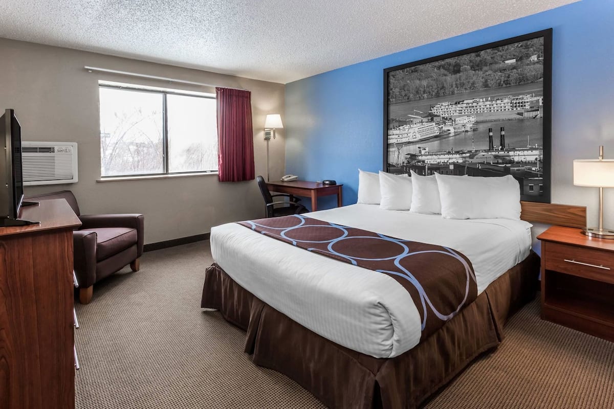 A hotel room with a large bed, a desk, a chair, a TV, a window with curtains, and a wall-mounted lamp.