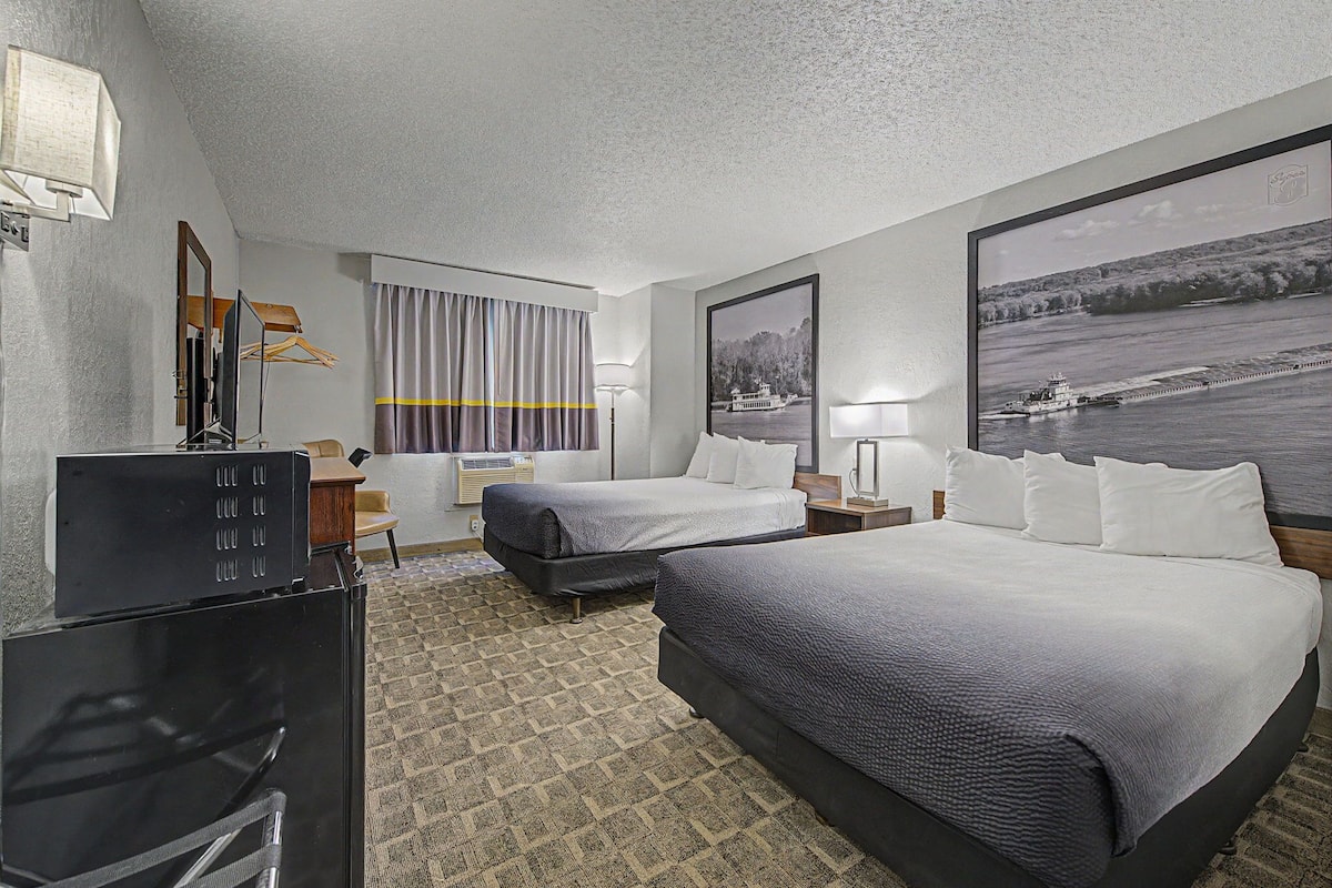 A hotel room with two beds, a desk, a TV, and a large window with curtains.