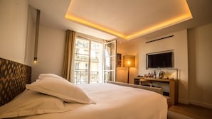 A hotel room with a bed, a desk with a TV, a lamp, and a view of the city. - Hotel Marignan Champs - Elysées (Paris)