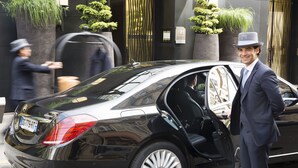 Un coche de lujo negro estacionado frente a un edificio de diseño moderno, un hombre con traje y sombrero de pie junto a él, y otro hombre caminando al paso.