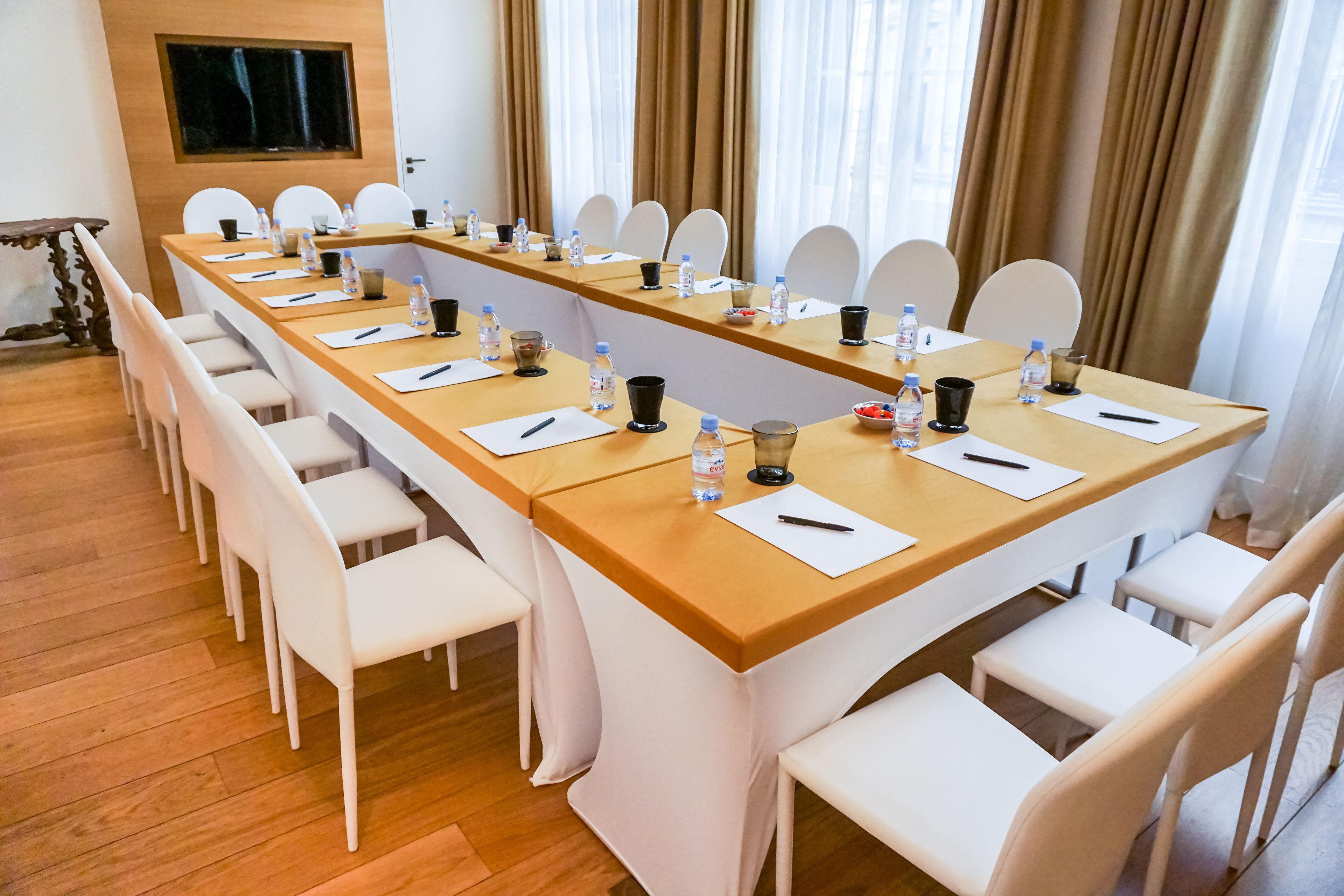 Una sala de conferencias con una larga mesa, sillas blancas y diversos elementos como botellas de agua y vasos.