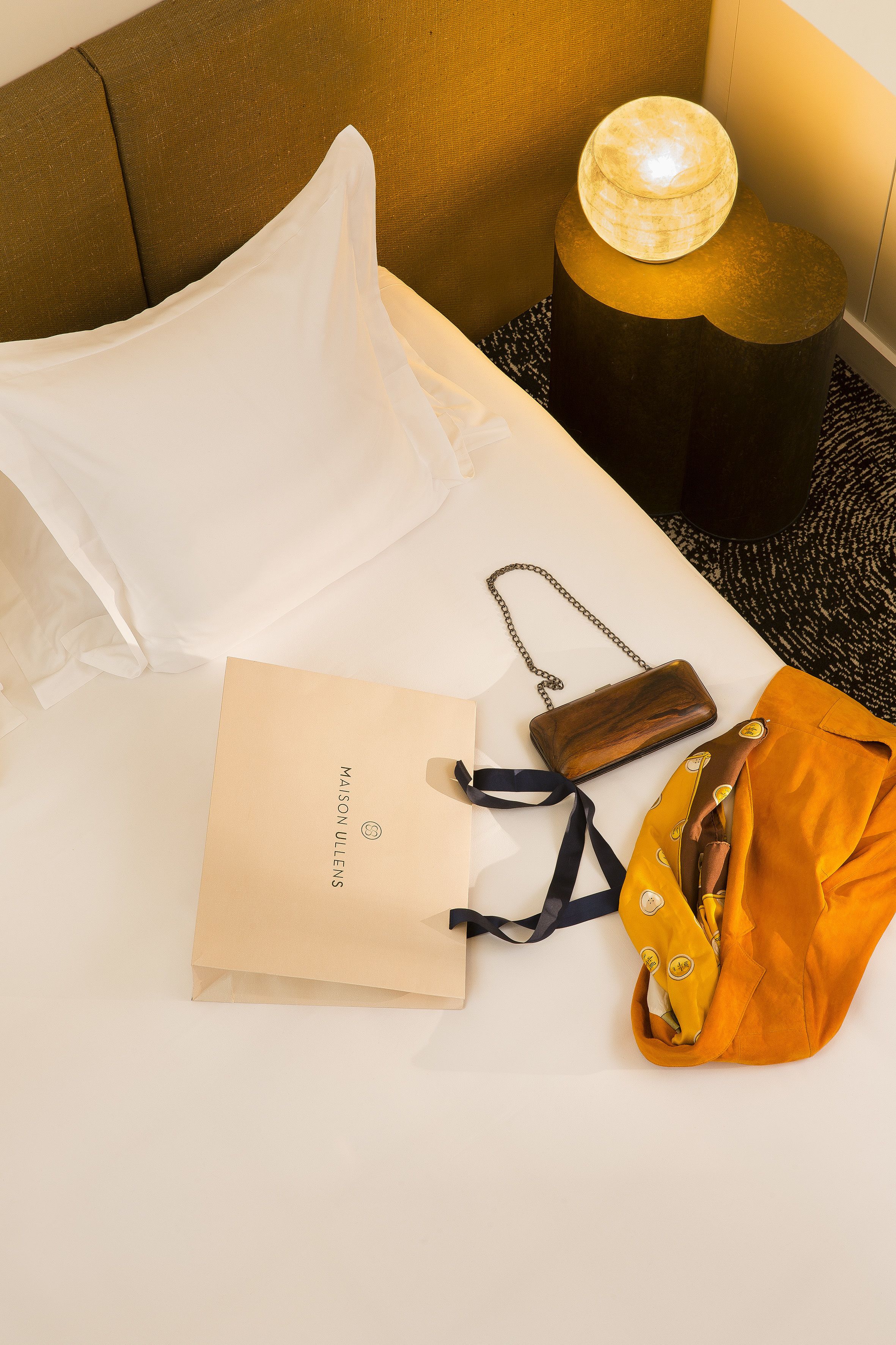 A neatly made bed with a white pillow, a brown bag, a yellow bag, and a brown paper bag with the text "MAISON ULLENS" on a white surface.