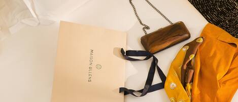 A neatly made bed with a white pillow, a brown bag, a yellow bag, and a brown paper bag with the text "MAISON ULLENS" on a white surface.