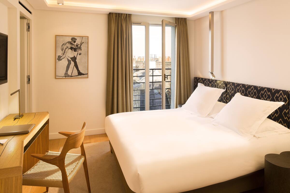 Habitación de hotel con cama, escritorio, silla, televisión y balcón con cortinas.