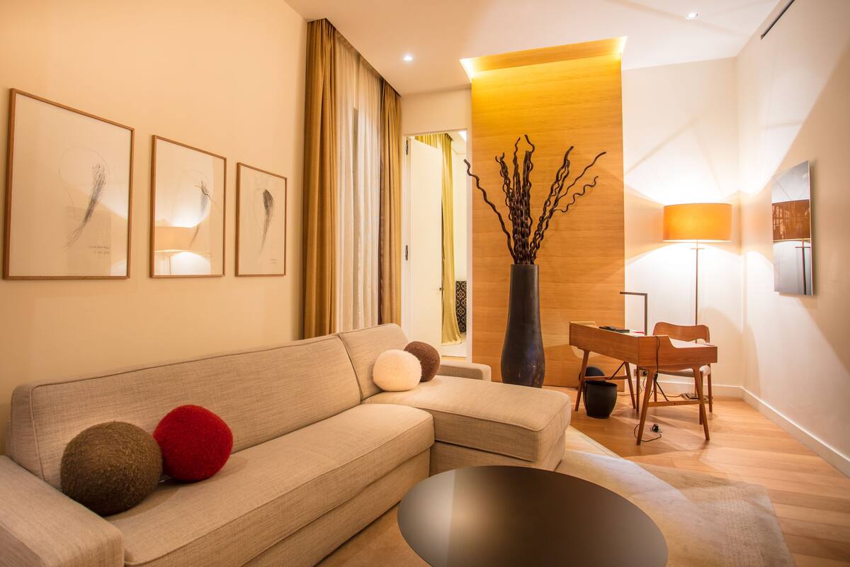 Una sala moderna con un sofá, una mesa de centro y una lámpara. Hay obras enmarcadas en la pared y un jarrón con ramas.