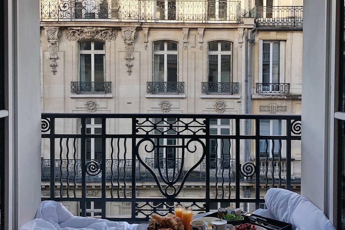 Un balcón con una bandeja de desayuno, una cama y vistas a un edificio clásico con balcones ornamentados.